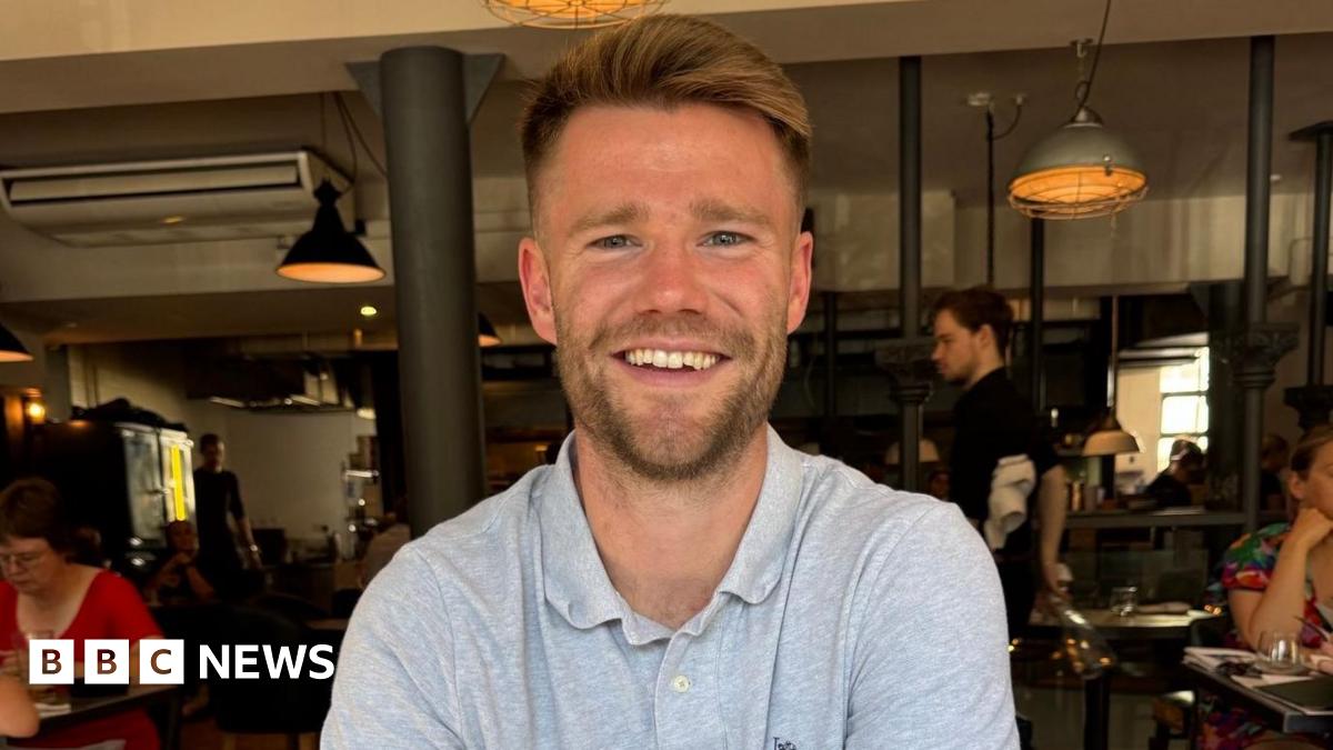 Hamish Douglas, a bearded man in his late 20s or early 30s smiling and wearing a grey polo shirt. He is sat inside a bar or restaurant, with industrial-style lighting behind him. Other customers can be seen at tables nearby.