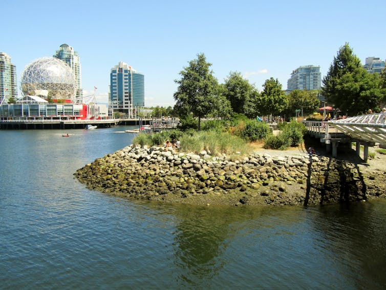 boulders along a shoreline near some trees. Buildings are seen in the background