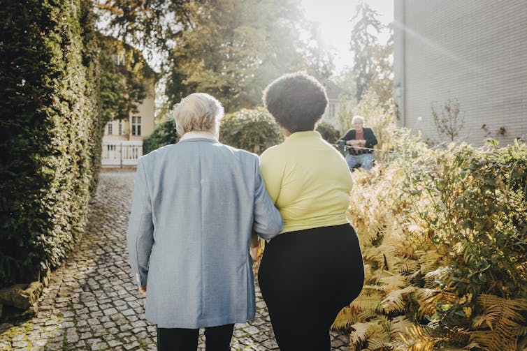 Rear view of female caregiver walking with retired male on footpath.