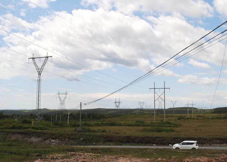 A car on a road driving past transmission towers and powerlines