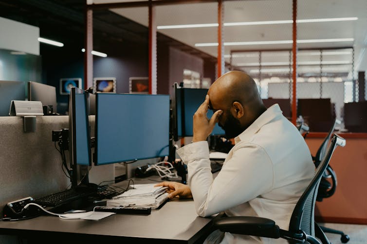 A man sits at a computer desk with his head in his hands