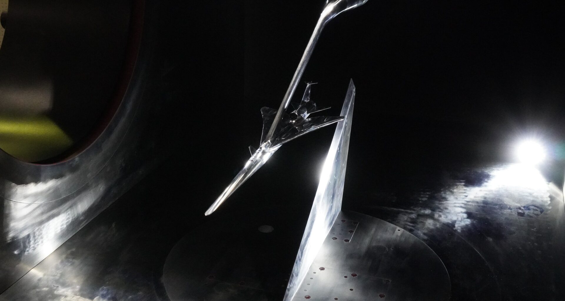 a long, narrow, silver model of an aircraft hangs from a boom in a dark concrete tunnel