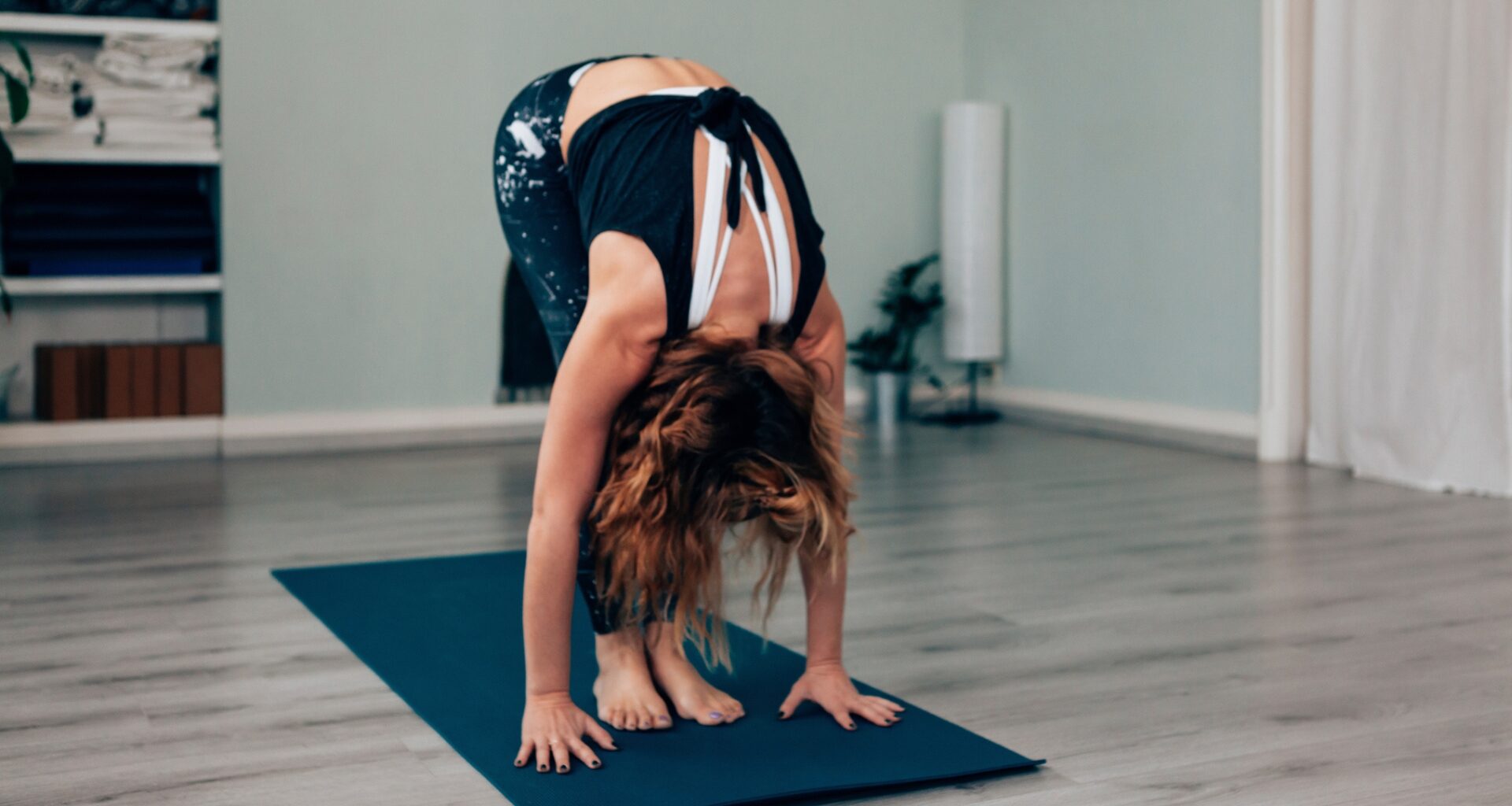 woman performing a rolldown from standing, touching her hands to the floor on an exercise mat and wooden floor.