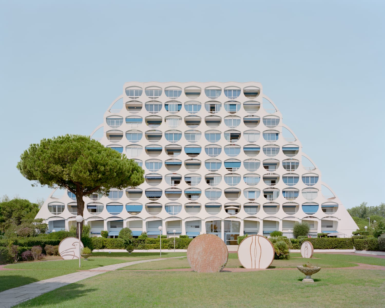 a modernist resort apartment complex in all-white against a blue sky with trees and sculptures in the foreground