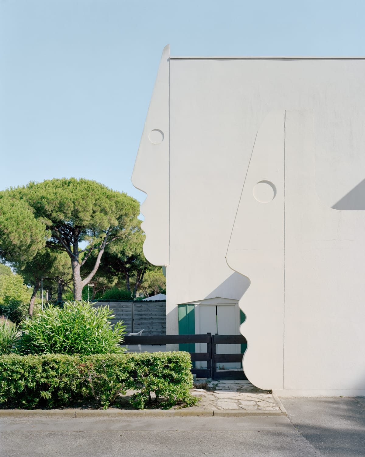 a detail of modernist architecture against a blue sky with shrubs, with shapes in the form of human facial profiles