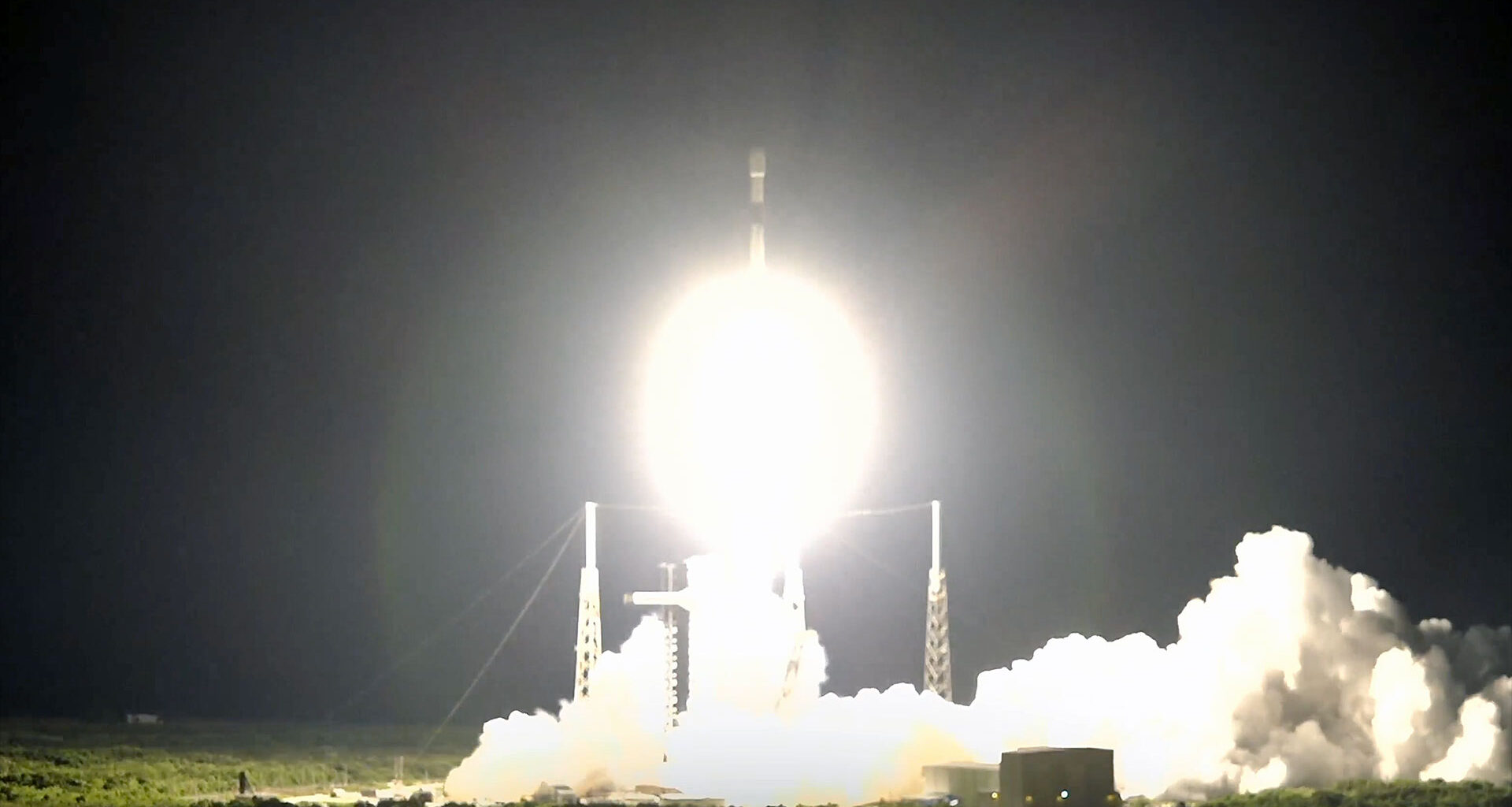 a white and black rocket launches into the dark of night, its bright white thrust lighting up its launch pad and surrounding area.