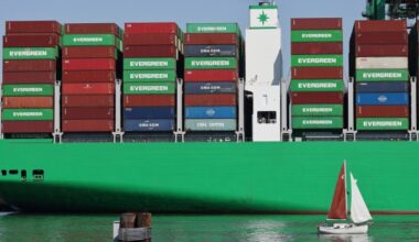 A sailboat passes shipping containers stacked on a container ship at the Port of Los Angeles