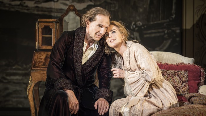 A woman in a pale robe leans against a man in a beaded velvet dressing gown