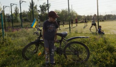A boy stands beside his bicycle adorned with a small Ukrainian flag near a public football field. In the background, several people are playing football on a grassy field enclosed by a wire fence
