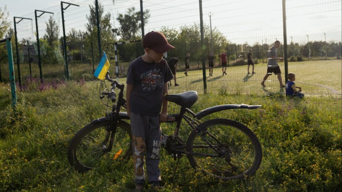 A boy stands beside his bicycle adorned with a small Ukrainian flag near a public football field. In the background, several people are playing football on a grassy field enclosed by a wire fence