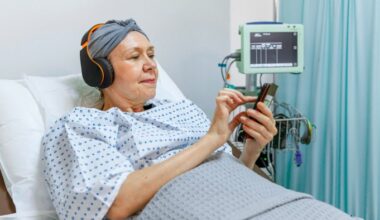 A woman with cancer who is wearing a scarf to hide hair loss from chemotherapy treatment lies in a hospital bed while wearing headphones and using a smartphone