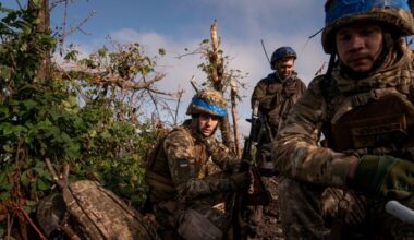 Soldiers wearing battle fatigues and helmets sit amid foliage, looking weary
