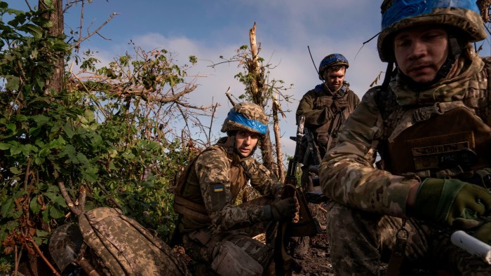 Soldiers wearing battle fatigues and helmets sit amid foliage, looking weary