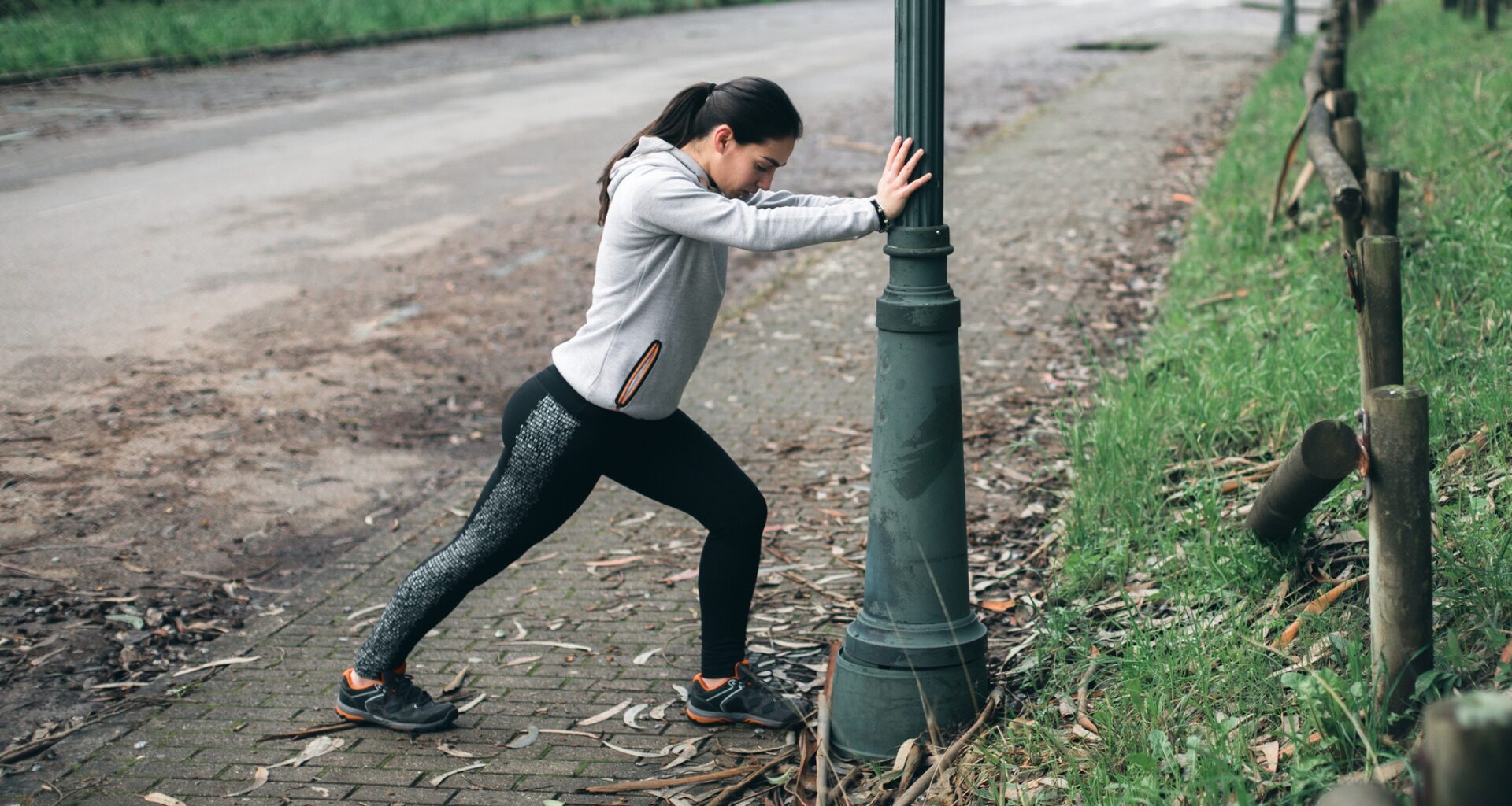 woman in leggings and hoodie doing a calf stretch against a lampost in a park setting
