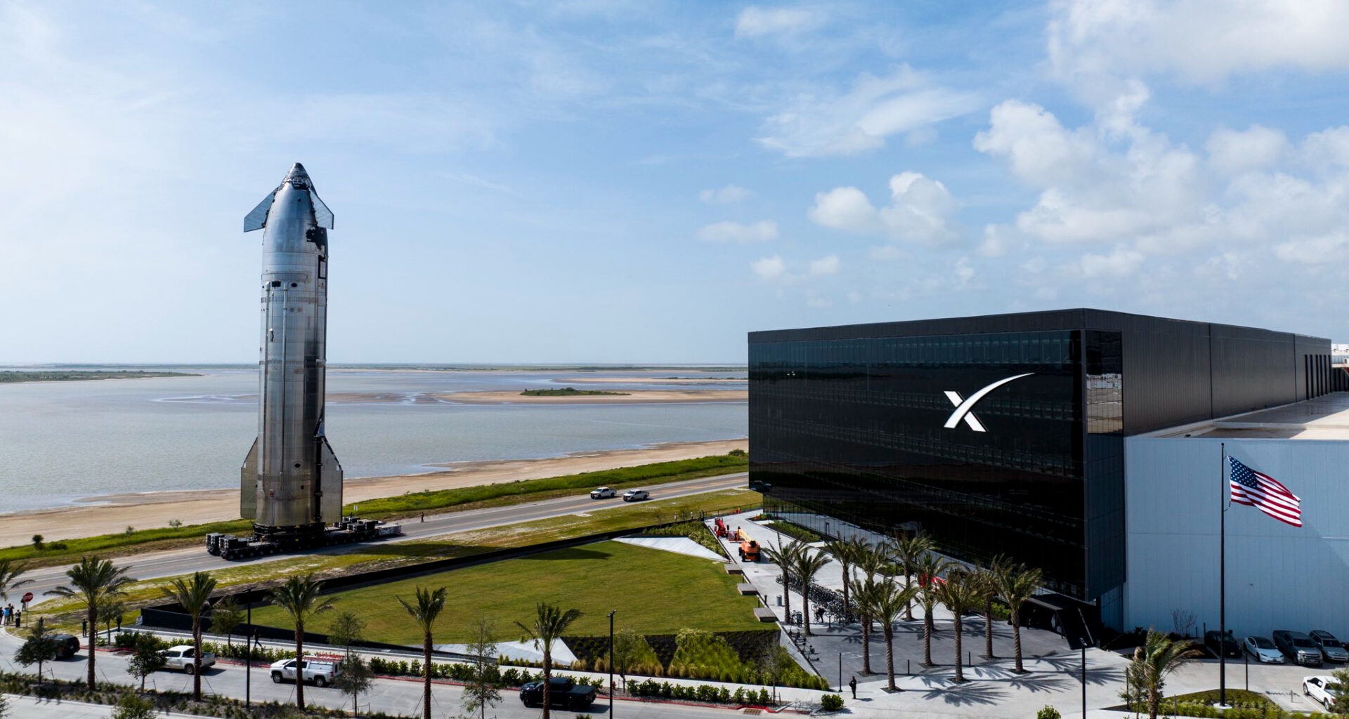 a large silver spacecraft rolls down a seaside road next to a large black building