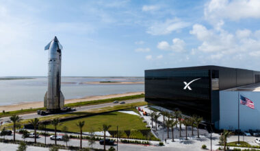 a large silver spacecraft rolls down a seaside road next to a large black building