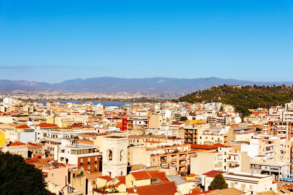 Aerial view of Cagliari, Sardinia, showing the city's buildings, Montelargius lake, and mountains in the background.
