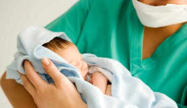 Image of a clinician holding a baby. The clinician is wearing green scrubs and is wearing a mask.