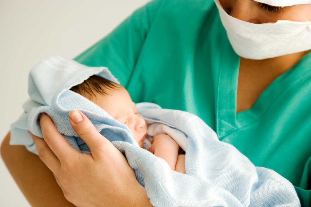 Image of a clinician holding a baby. The clinician is wearing green scrubs and is wearing a mask.