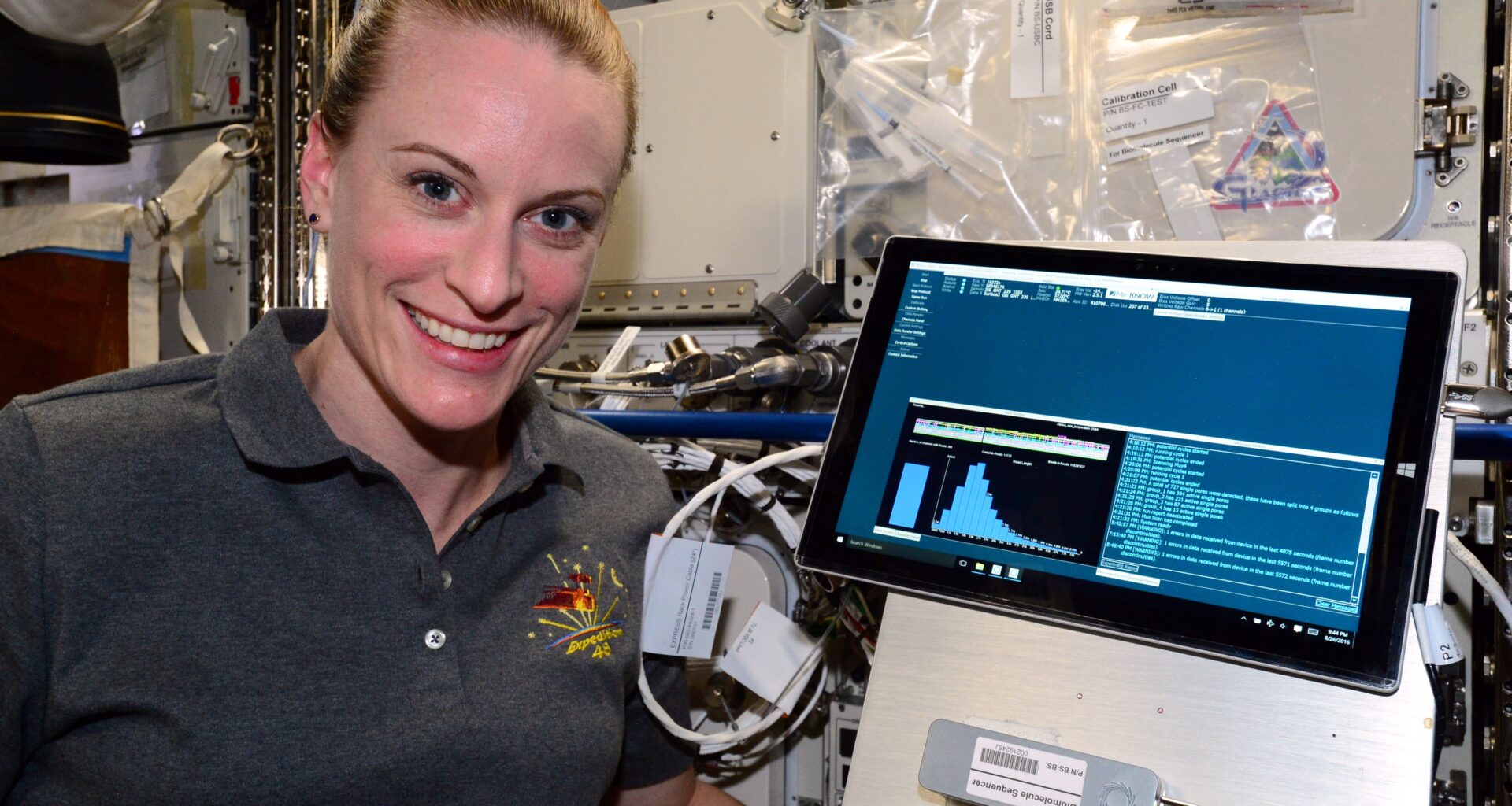 headshot of a smiling woman in a gray polo shirt in a cluttered laboratory setting, with a computer monitor next to her