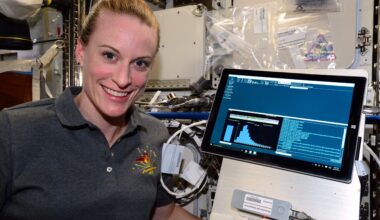 headshot of a smiling woman in a gray polo shirt in a cluttered laboratory setting, with a computer monitor next to her
