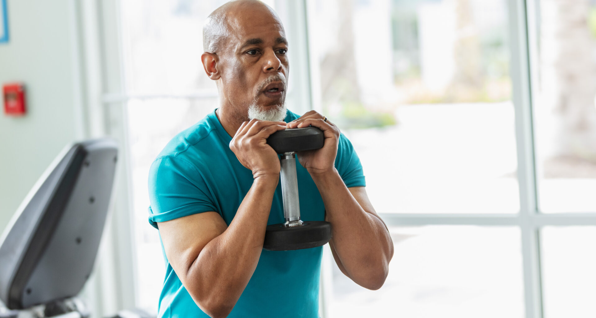 A man in a t-shirt holds a dumbbell to his chest. Behind him we see the rear of a screen attached to an exercise bike.