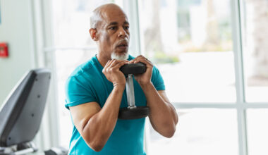 A man in a t-shirt holds a dumbbell to his chest. Behind him we see the rear of a screen attached to an exercise bike.