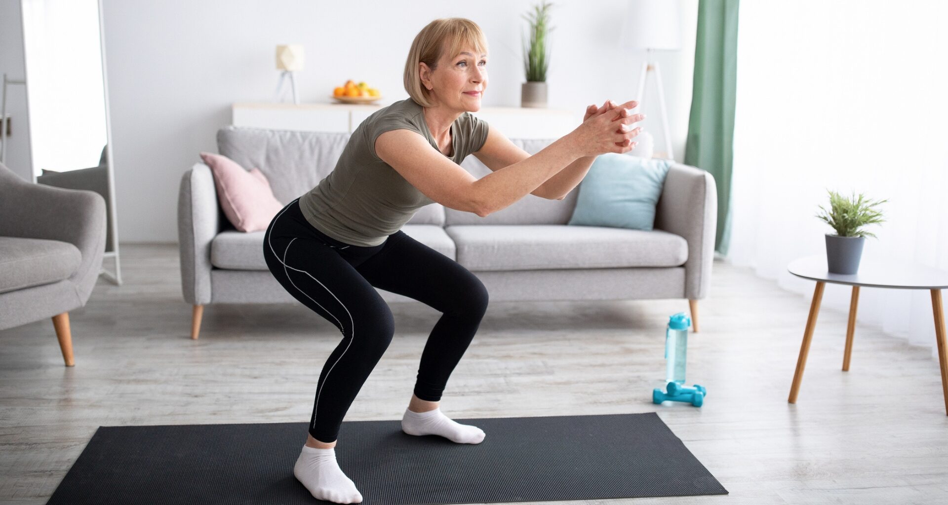 Woman performing a shallow squat on a black exercise mat in a living room setting. there's a sofa behind her.