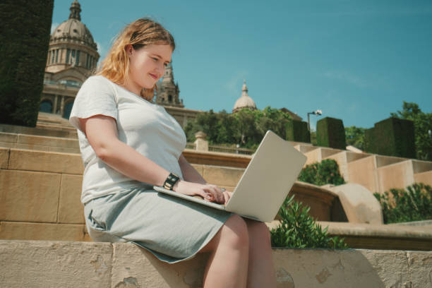 Young woman uses a laptop while sitting on steps near a historic building with domes and greenery.