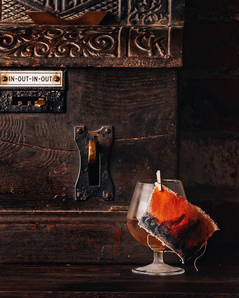 Cocktail glass with painted fabric garnish next to antique wooden box.