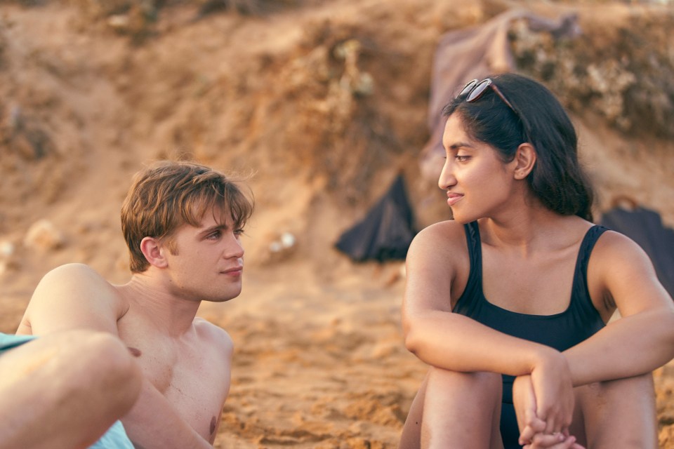 A young man and woman sitting on a beach, looking at each other.