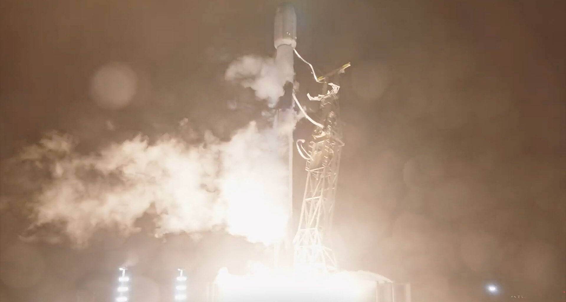 a tall white rocket lifts off into the night sky, its bright white engine plume lighting the area below