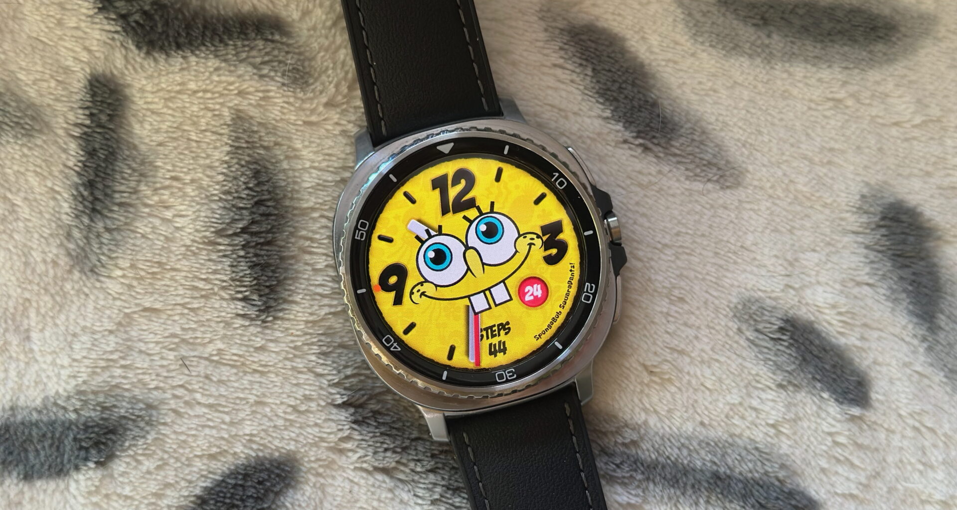 The Samsung Galaxy Watch 8 Classic resting on a blanket showing a custom Facer watch face of Spongebob Squarepants.