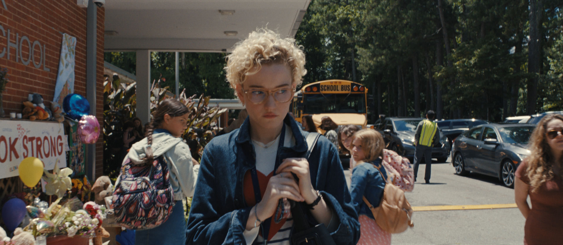 A scene outside a school showing a young woman with curly hair and glasses, looking somber while holding a bag. The background features students and a yellow school bus, with a memorial display reading "LOOK STRONG".