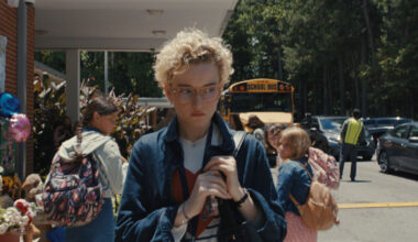 A scene outside a school showing a young woman with curly hair and glasses, looking somber while holding a bag. The background features students and a yellow school bus, with a memorial display reading "LOOK STRONG".