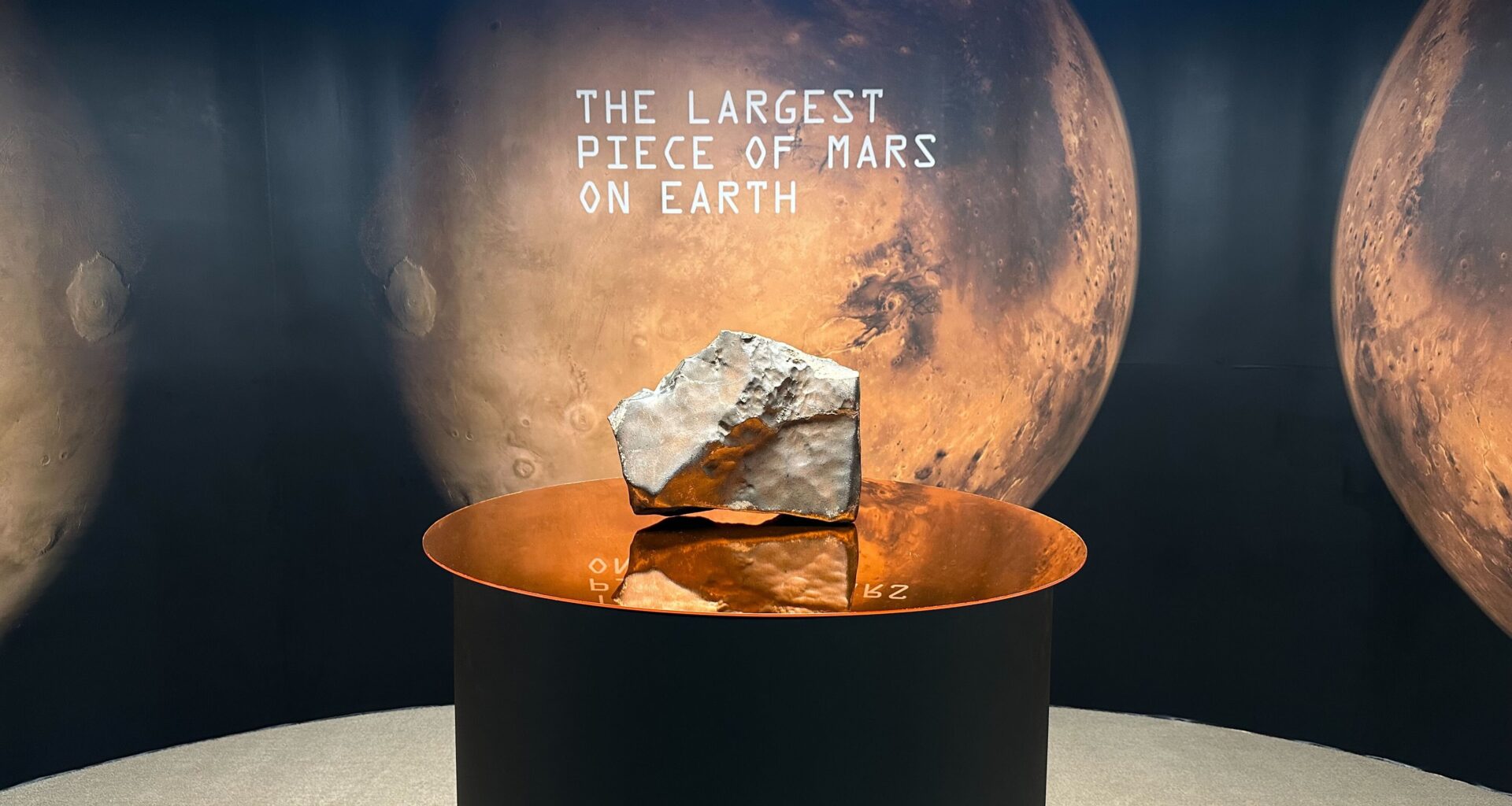 A large rock on a pedestal with a sign in the background that has a picture of Mars and says &quot;The Largest Piece of Mars On Earth.&quot;