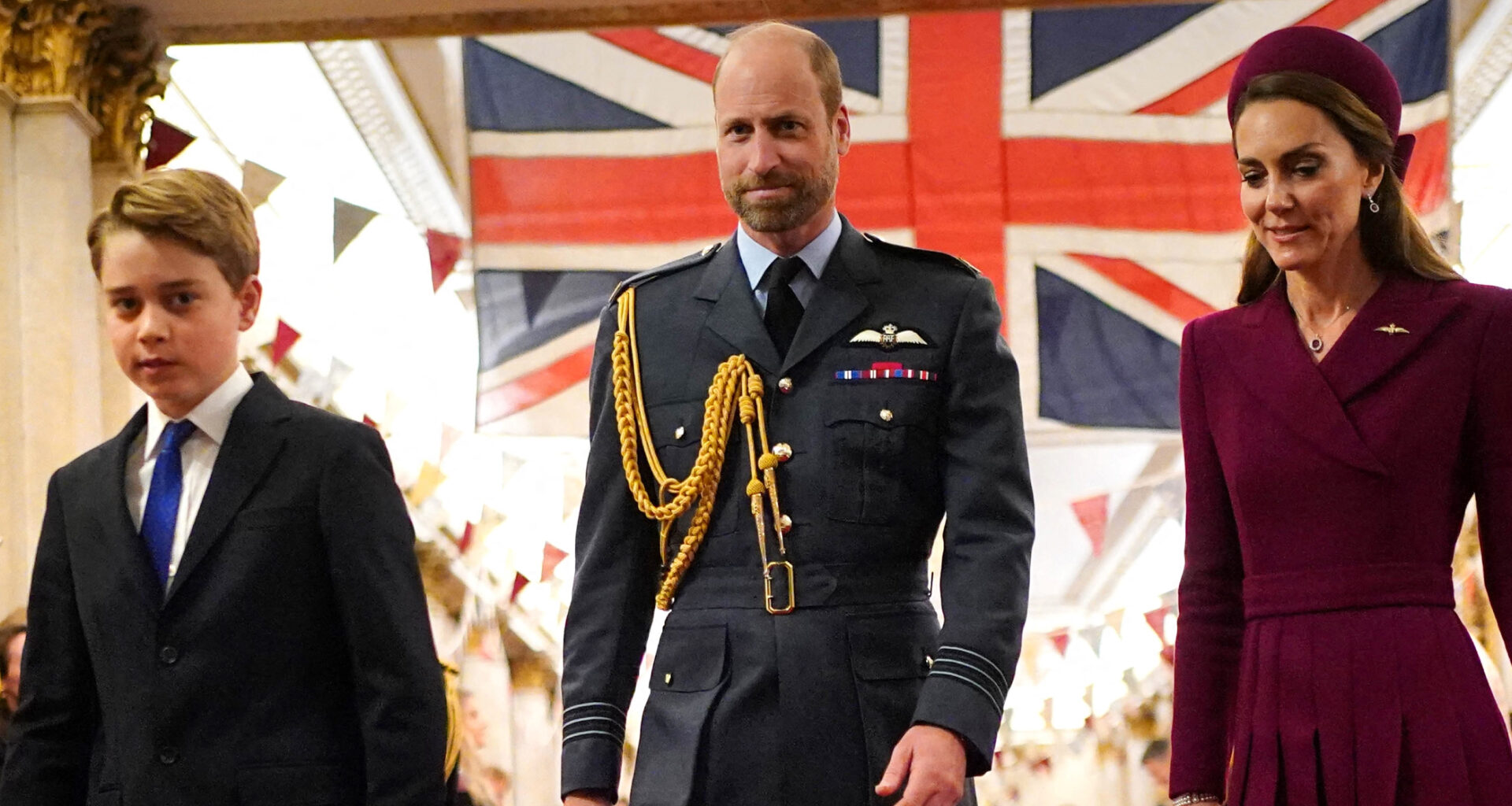 Prince George wears a suit, Prince William wears military uniform, Kate Middleton wears a dark red coat dress, as they walk in front of a Union Jack flag