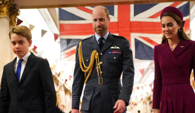 Prince George wears a suit, Prince William wears military uniform, Kate Middleton wears a dark red coat dress, as they walk in front of a Union Jack flag