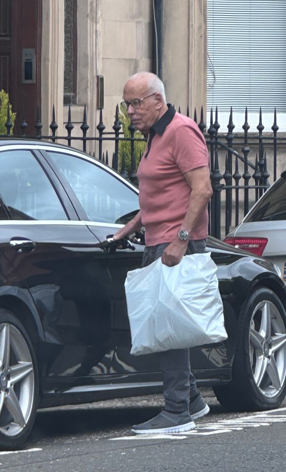 An older man with glasses gets into a black car, carrying a large plastic bag.