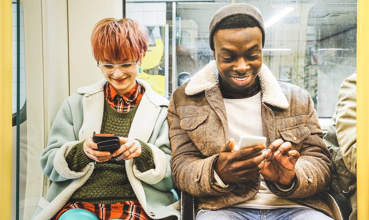 A multiracial duo sit in a well-lit London Underground tube train, both smiling and playing on their phones.