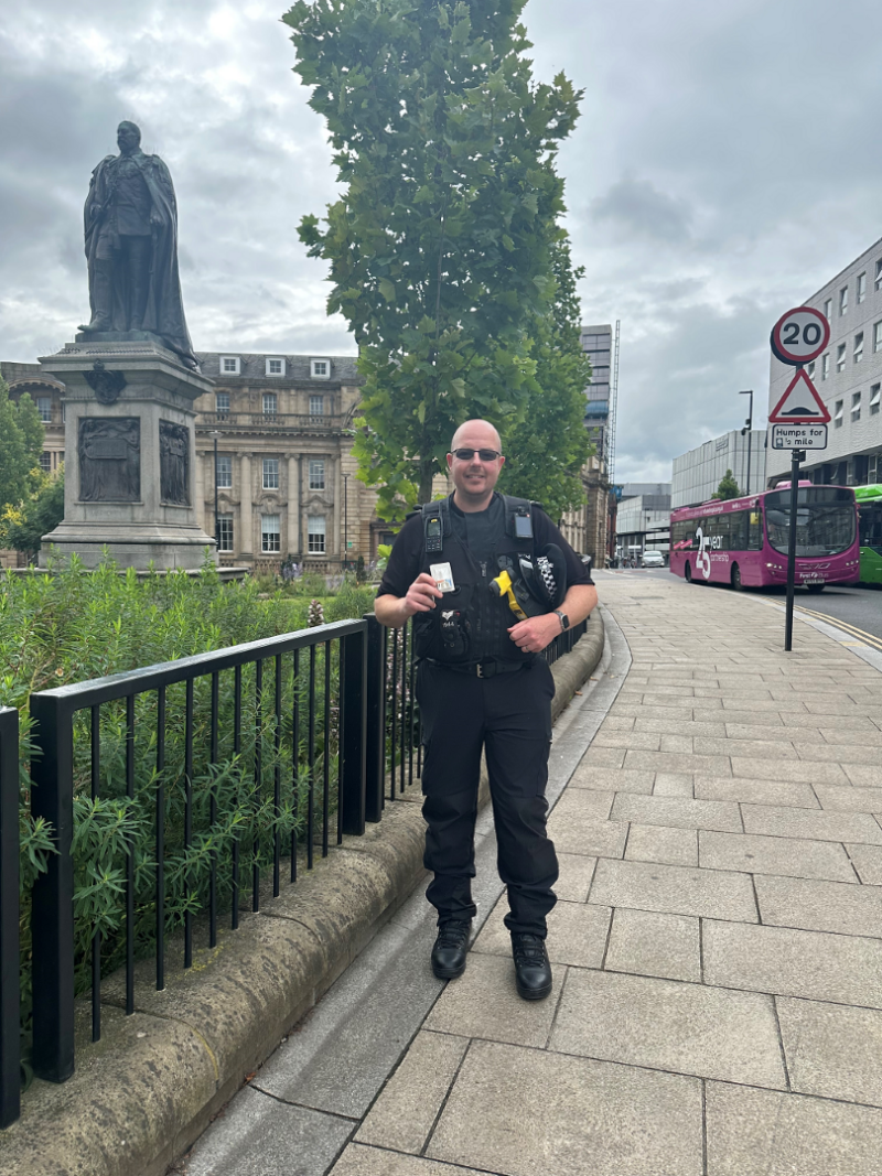 A photo of a smiling police officer standing in the street, wearing sunglasses and holding up a box of a medicine