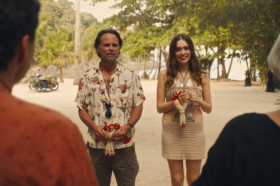 Scene from The White Lotus season 3: a man and woman holding flower leis on a beach.