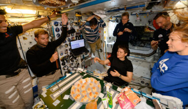 seven people in polo shirts and slacks gather around a table to enjoy a meal together aboard a space station