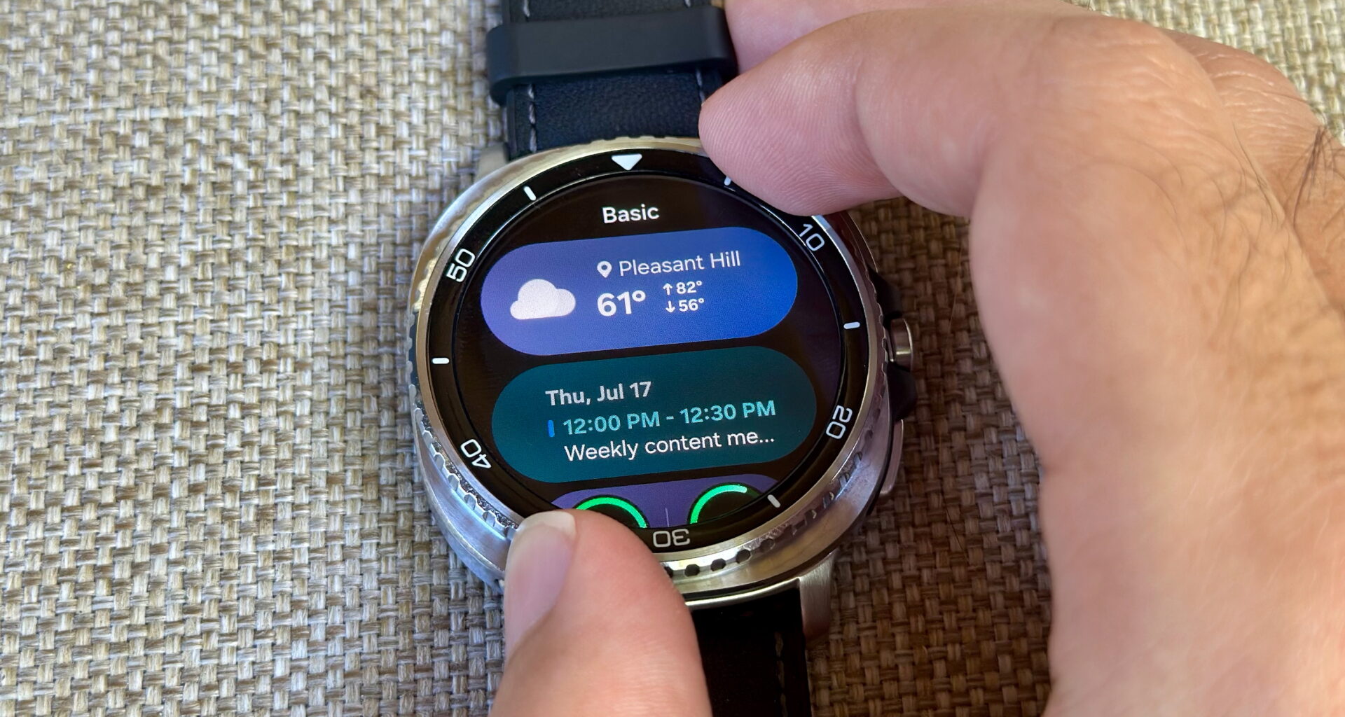 Two fingers gripping the Samsung Galaxy Watch 8 Classic rotating bezel, the display turned to a Tile with weather and calendar data