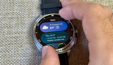 Two fingers gripping the Samsung Galaxy Watch 8 Classic rotating bezel, the display turned to a Tile with weather and calendar data