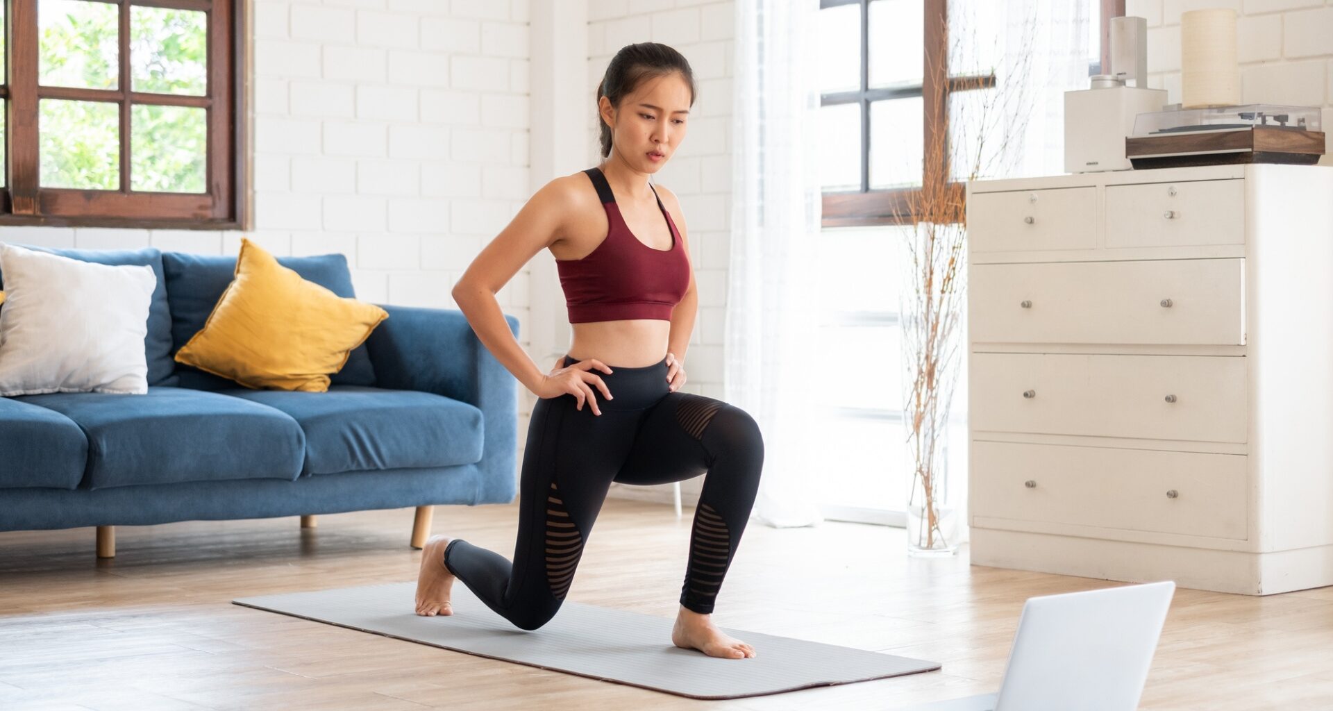 woman in black leggings and red crop top performing a lunge on an exercise mat in front of a laptop on wooden floor. there's a navy sofa and windows behind her and a white chest of drawers next to her.