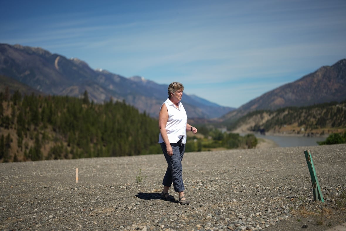 A woman in a white, sleeveless shirt and blue pants walks on a patch of gravel, framed by mountains, trees and a body of water behind her. 