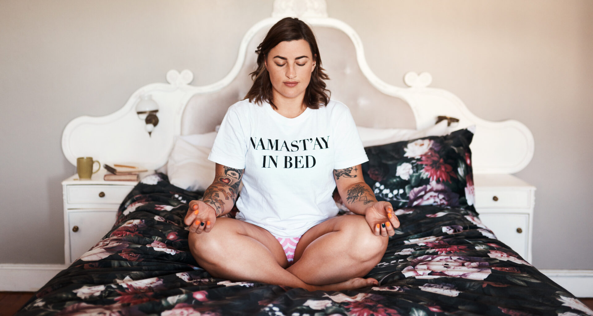 Woman sits cross-legged on a bed spead wearing a white T-shirt that reads NAMAST’AY IN BED
