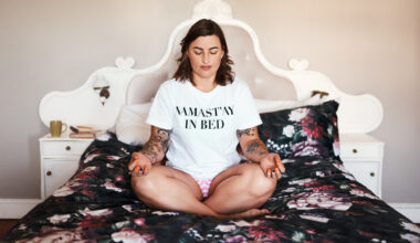Woman sits cross-legged on a bed spead wearing a white T-shirt that reads NAMAST’AY IN BED