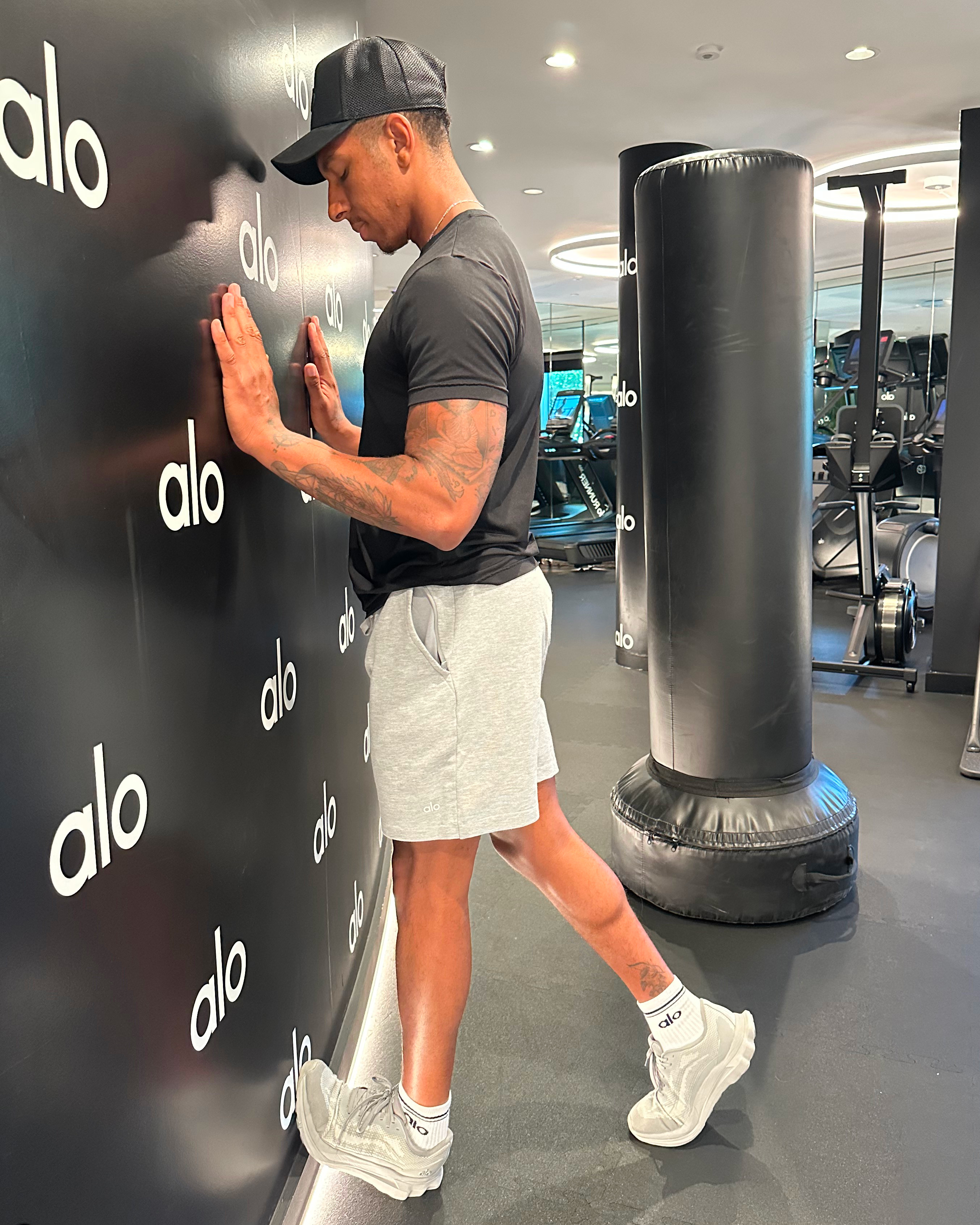 Man in black T-shirt, black cap, white shorts and white trainers demonstrates exercise
